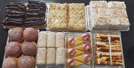 Surtido de pasteles y postres en bandejas metálicas, incluyendo eclairs con chocolate, tartas de banana, pasteles con crema y coco, porciones de cheesecake decoradas y postres con merengue tostado.